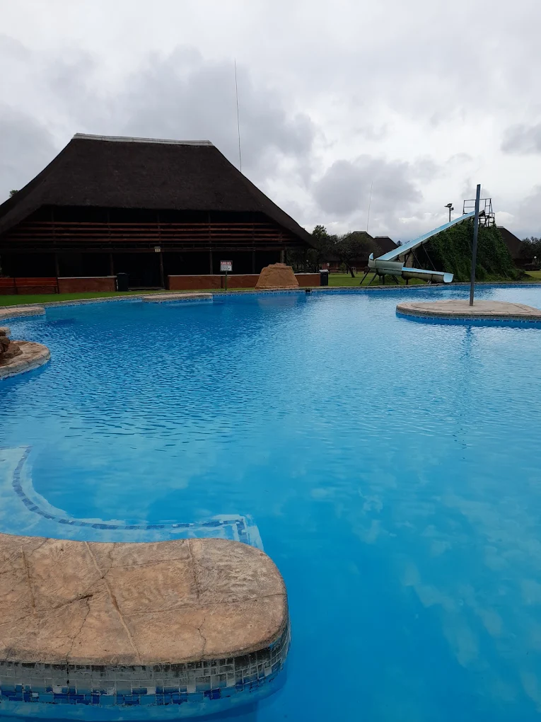 Freeform pool with stone ledges, a thatch hall and a slide on a grassy bank